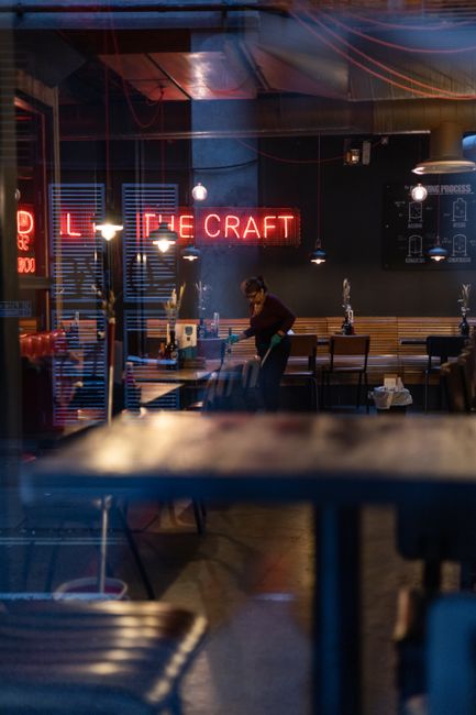 A worker wearing yellow rubber boots and casual clothing is engaged in surface cleaning outside a retail shop in Shoreditch, London. The worker uses a power washer to clean the pavement area, creating