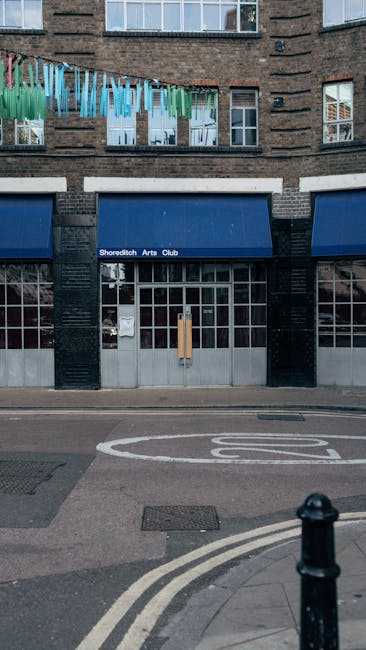 A street scene in Shoreditch N1 showing modern glass office buildings and older brick structures with a large graffiti-covered billboard sign in the foreground. The street includes signage for bus sto